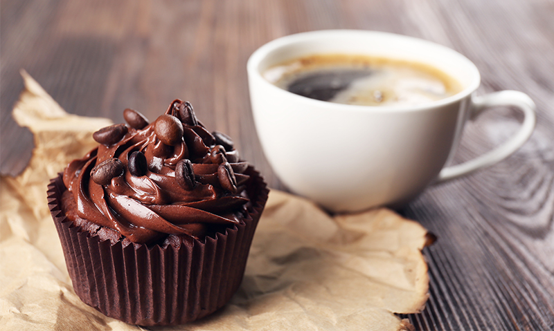 Chocolate cupcake with glossy chocolate frosting and coffee cup on a wooden table.