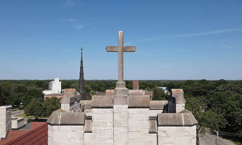 Stone cross atop a church roof with a spire and treeline under a clear blue sky.